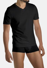 Uomo che indossa una T-shirt nera a V e boxer neri, in piedi contro uno sfondo chiaro uniforme, testa parzialmente fuori dal fotogramma.