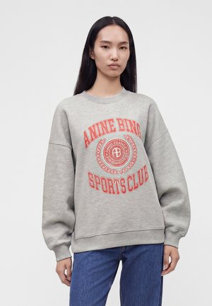 Jeune femme aux longs cheveux noirs portant un sweat gris avec un texte rouge "ANINE BING SPORTS CLUB" et un jean bleu, debout devant un fond uni.