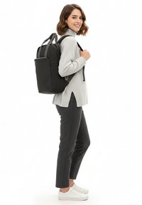 Musta reppu, joka on valmistettu sileästä materiaalista, kahdella olkahihnalla, kapealla suorakaiteen muotoisella rakenteella ja etutaskulla. Malli pukeutuneena harmaaseen neulepaitaan ja housuihin.