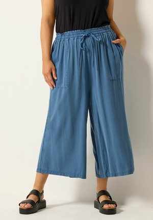 Kobieta ubrana w luźne, niebieskie spodnie o wysokim stanie, skrócone, z przednimi kieszeniami, sznurkiem, czarne sandały i czarny top bez rękawów.