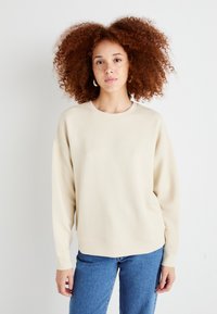 Beige Sweatshirt mit Rundhalsausschnitt, überschnittenen Schultern und gerippten Bündchen. Kombiniert mit blauen Jeans. Weicher Stoff, schlichtes Design.
