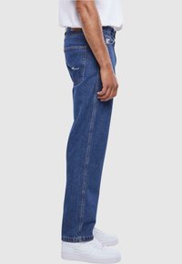 Blå denimjeans med rett ben design, med detalj på baklommen og en synlig midjebåndsetikett. Kombinert med hvite sneakers.