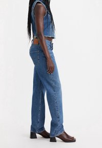 Jeans de mezclilla de pierna amplia, de color azul claro con dobladillo deshilachado, con una etiqueta de cuero marrón en la parte posterior y un corte holgado. Combinados con sandalias de tacón.