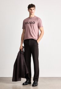 Ružové tričko s nápisom "HUGO" potlačeným čierne, skombinované s tmavými nohavicami. Model drží tmavohnedú bundu a má na sebe čierne topánky s hladkým povrchom.