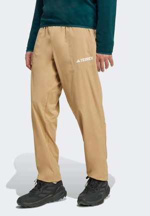 Pantalons légers beiges avec une taille élastique, dotés de poches latérales et d'un logo "TERREX" blanc à l'avant, associés à des chaussures noires.