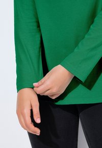 Zielona koszula z długim rękawem o gładkiej fakturze i zaokrąglonym dekoltem, z bocznymi rozcięciami i dopasowanymi mankietami. Czarne spodnie widoczne częściowo.