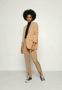 Mulher com penteado afro a usar um fato bege, suéter preto de gola alta e ténis pretos, de pé contra um fundo claro e simples.