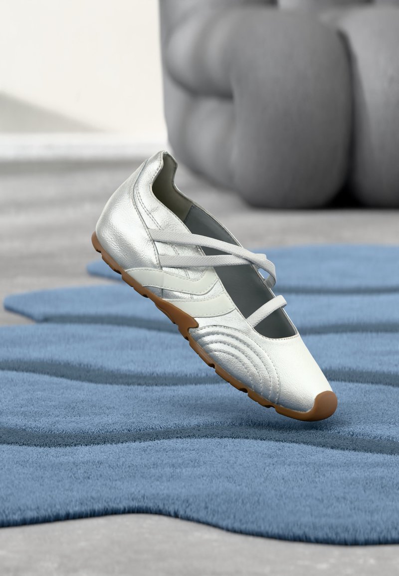 Chaussure de sport blanche et argentée avec une semelle marron flottant au-dessus d'un tapis bleu texturé dans un décor intérieur minimaliste.