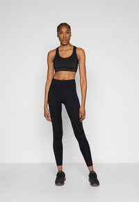 Czarny sportowy biustonosz i legginsy z wysokim stanem wykonane z elastycznego materiału. Modelka stoi w czarnych sneakersach. Gładka tekstura, dopasowany fason.