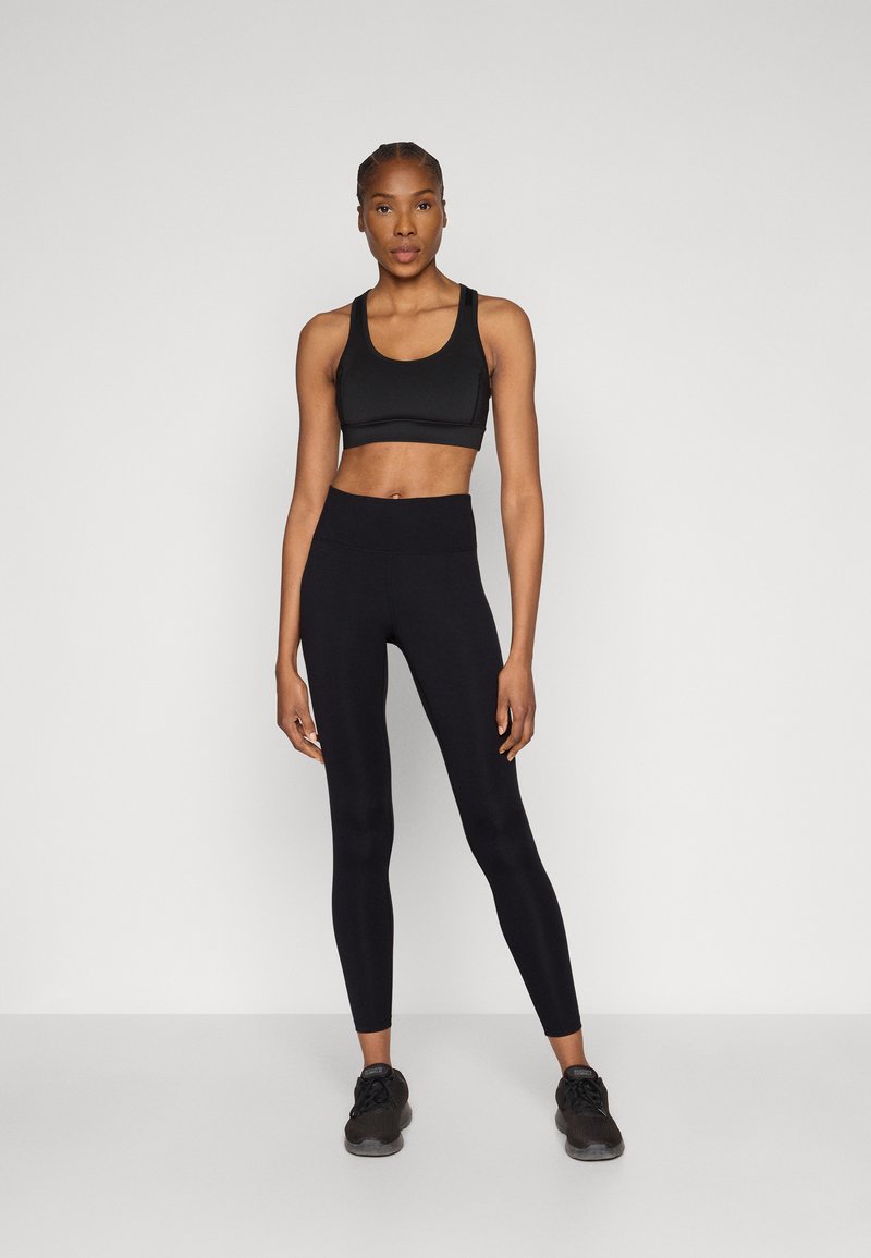 Melns sporta krūšturis un augstas jostasvietas legingi no elastīga auduma. Modele stāv melnās sporta kurpēs. Gluda tekstūra, pieguļošs dizains.