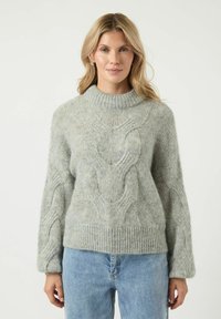Jasnoszary sweter z dzianiny z wysokim kołnierzem, z wzorem w warkocze i ściągaczem u dołu. Luźny krój z długimi, szerokimi rękawami. W zestawie z niebieskimi dżinsami.
