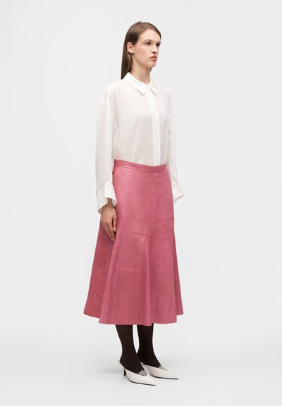 Φούστα από ροζ δέρμα με φουσκωτή βάση, συνοδευόμενη από λευκό διαφανές πουκάμισο με κουμπιά και μικρές υφασμάτινες κουκκίδες, καθώς και λευκά παπούτσια με τακούνι.