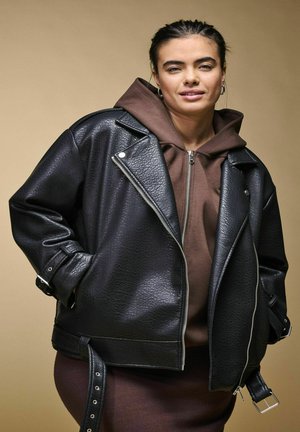 Jeune femme aux cheveux foncés porte des boucles d’oreilles anneaux, un sweat à capuche marron et une veste en cuir noire texturée avec des fermetures éclair et une ceinture.