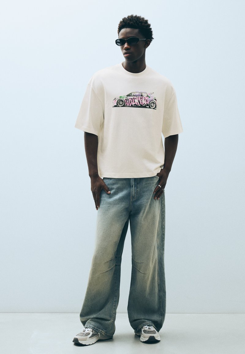Jeune homme portant un t-shirt blanc oversize avec un imprimé voiture coloré, un jean large bleu délavé, des baskets et des lunettes de soleil noires, debout sur un fond uni.