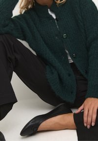 Cardigan di un verde scuro con texture e colletto alto, dotato di bottoni neri e una vestibilità ampia, abbinato a pantaloni neri e scarpe basse nere.