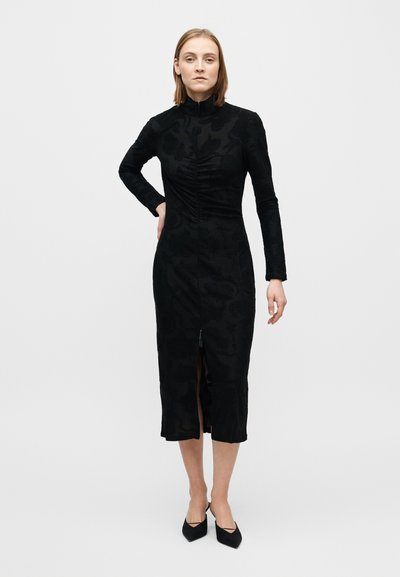 Fekete ruha hosszú ujjakkal, magas nyakkal és elején ráncolt díszítéssel. Elöl hasítékkal és texturált virágmintával rendelkezik. Középhosszúságú, a borjú közepéig ér.