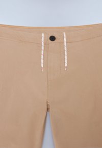 Beige shorts med en glat tekstur, prydet med en sort knap, hvid snor med orange detaljer og en subtil syet sømdetail.