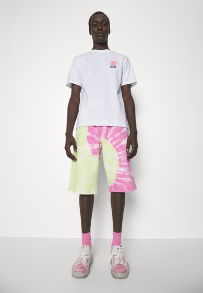 Fehér, rövid ujjú póló, kis logóval, amelyet rózsaszín és lime zöld batikolt szorttal párosítanak. Rózsaszín zoknikat és vastag talpú sportcipőt visel.