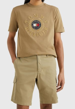 Mies pukeutunut beigeen Tommy Hilfiger -logolliseen t-paitaan ja siihen sopiviin beigen sävyisiin cargo-shortseihin, kädet taskuissa, yksivärisen harmaan taustan edessä.