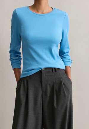 Person trägt ein eng anliegendes hellblaues Langarmshirt kombiniert mit hoch taillierten, weiten dunkelgrauen Hosen mit den Händen in den Taschen.