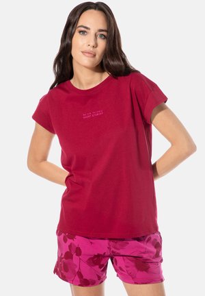 Frau mit langem dunklem Haar, die ein rotes T-Shirt mit der Aufschrift "No risk, no story. SHORT STORIES" und rosa geblümte Shorts trägt, steht mit den Händen hinter dem Rücken.