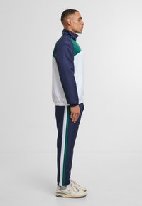 Tuta della marina con accenti bianchi e verdi; la giacca ha un colletto alto e polsini elasticizzati; i pantaloni presentano strisce laterali; abbinati a sneakers beige.