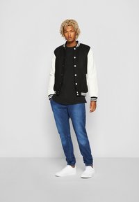 Chaqueta varsity en blanco y negro con puños a rayas, llevada sobre una camiseta negra, combinada con jeans azul entallados y zapatillas blancas.