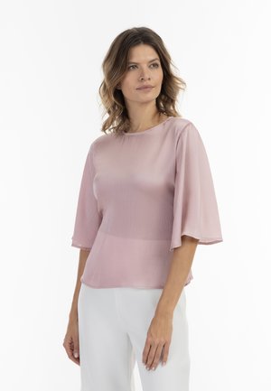 Blusa rosa trasparente con maniche corte e fluide e scollatura rotonda. Il tessuto presenta un motivo testurizzato sottile. Abbinata a pantaloni bianchi.
