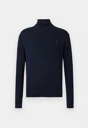 Navy ribbed knit sweater with long sleeves, high collar, four black buttons, and a small red, white, and blue logo on the chest.