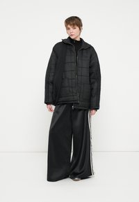 Casaco acolchoado preto com capuz alto, fecho de zip e bolsos laterais, combinado com calças largas pretas de cetim com riscas brancas laterais.