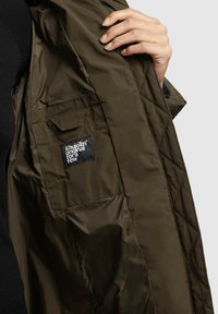 Oliivivihreä tikattu takki, jonka pinta on sileä. Varustettu erillisellä taskulla ja sisäpuolella brändilogo, mikä korostaa yksinkertaista muotoilua.