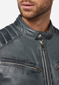 Grijze leren jas met een gestructureerde afwerking, hoge kraag en zichtbare stiksels. Voorzien van zilveren ritssluitingen aan de voorkant en op de bovenste zakken.