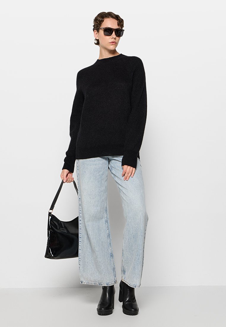 Schwarzer gerippter Strickpullover, hellblaue weit geschnittene Jeans, schwarze klobige Knöchelstiefel und eine schicke schwarze Handtasche; Model trägt dunkle Sonnenbrille.