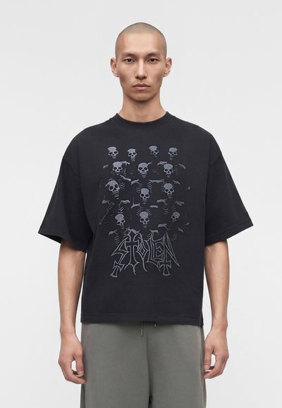 Mann trägt schwarzes oversized T-Shirt mit grauen Skelettgrafiken und dem Schriftzug "STOLEN", kombiniert mit grauer Hose vor neutralem Hintergrund.