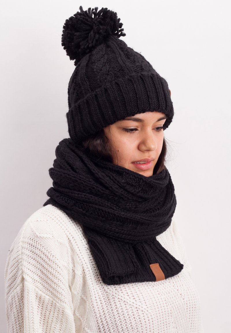 Black knitted beanie with a pom-pom and matching scarf. Both feature a textured, ribbed design. Scarf includes a leather accent.