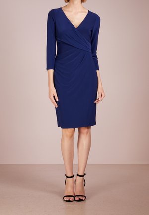 Kvinde iført en knælang, langærmet navyblå slå-om-kjole med sorte ankelstroppede hæle, stående foran en ensfarvet beige baggrund.