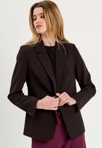 Blazer noir avec une coupe ajustée, des revers crantés, une fermeture à un bouton et des manches longues. Fabriqué en tissu lisse avec un design minimal.