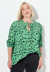 Grøn bluse med sorte polka-dots, knapbinding ved halsen, rynket tekstur og lange ærmer. Kantlinjen er asymmetrisk. Parret med sorte bukser.