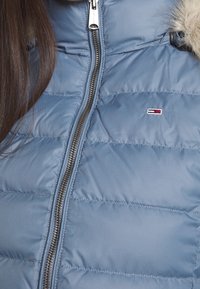 Zbliżenie osoby w jasnoniebieskiej pikowanej kurtce z srebrnym zamkiem błyskawicznym i małym czerwono-biało-niebieskim prostokątnym logo na piersi.