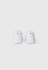 White sneakers with a smooth leather-like material, round toe, padded collar, and a textured rubber sole, featuring a small multicolored logo on the back.