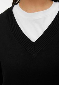 Czarny sweter z ryflowanym dekoltem w kształcie litery V noszony na białej koszulce. Miękka faktura, zbliżenie podkreślające dekolt i nałożenie warstw.