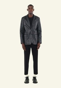 Zwarte textuurblazer met een enkele knoop, gecombineerd met een strakke zwarte broek en zwarte schoenen. Model staat tegen een lichte achtergrond.