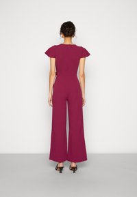 Une femme aux cheveux bouclés coiffés en un chignon bas porte une combinaison magenta avec des manches courtes et volantes et un pantalon large, associée à des sandales à talons noires.