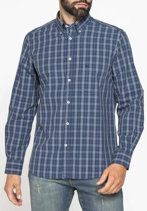 Uomo che indossa una camicia a quadri blu e bianca a maniche lunghe abbottonata, abbinata a jeans azzurri strappati.