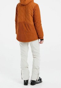 Braune Cordjacke mit rautenförmigem Steppmuster, Kapuze und elastischen Bündchen, kombiniert mit weißen Schneehosen und schwarzen Skischuhen.