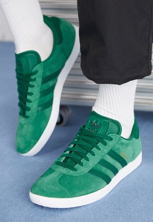 Grüne Wildledersneaker mit dunkelgrünen Streifen und weißen Sohlen, getragen mit weißen Socken und schwarzen Hosen auf blauem Boden.