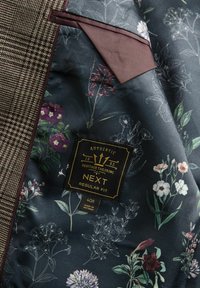 Gestreifter Blazer-Innenraum mit dunklem Stoff und floralen Mustern in Grün, Lila und Weiß. Karierter Taschenbereich und schwarzes Etikett mit goldener Schrift.