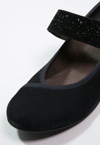 Ballet en cuir de daim noir avec un bout arrondi, présentant une sangle décorative noire avec des accents de perles et une doublure douce grise à l'intérieur.