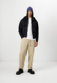 Svart zip-up hettegenser med frontlommer, hvit t-skjorte, beige vide bukser og svarte klumpete sko. Blå ribbet lue på hodet.