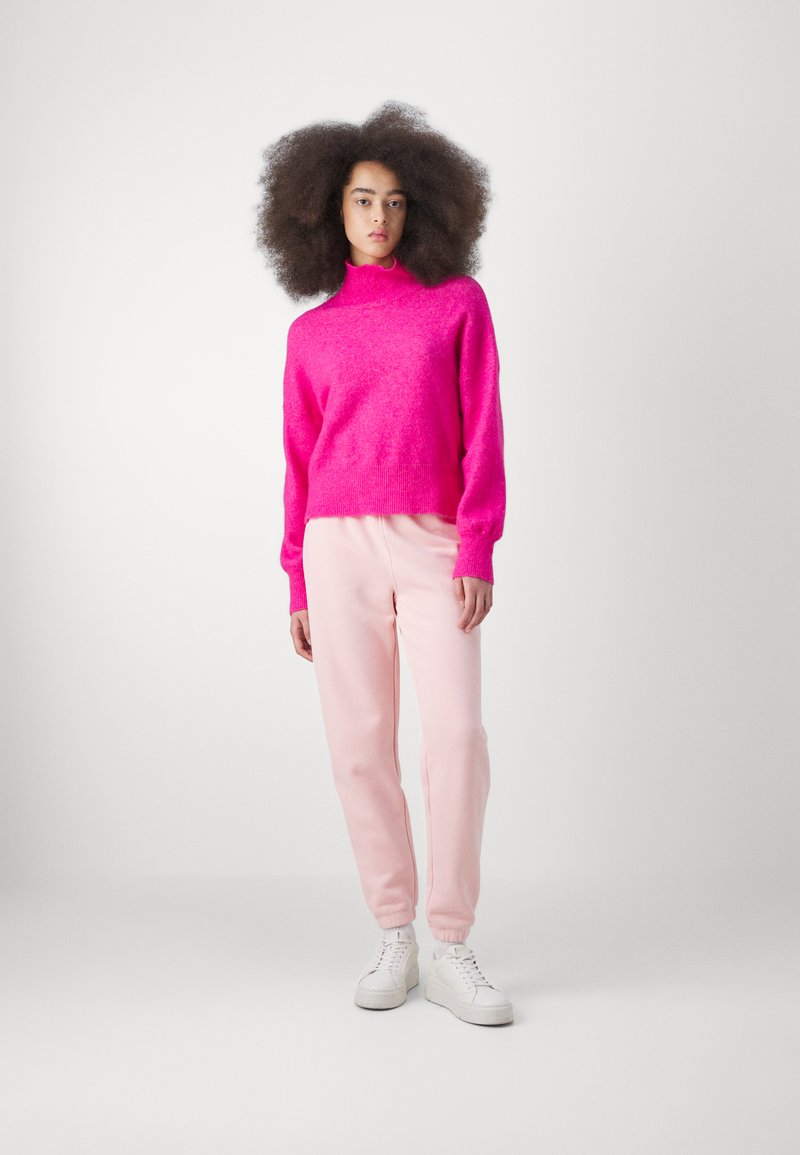 Pull en laine rose duveteux avec col montant, associé à un pantalon de survêtement rose clair et des baskets blanches ; le modèle se tient contre un fond blanc uni.